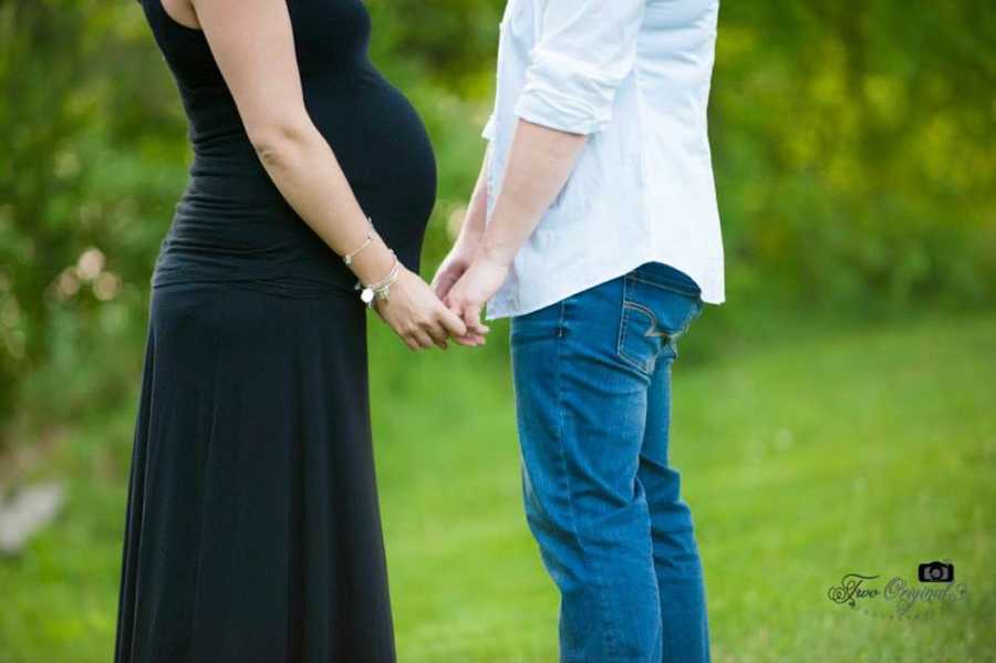 Married wives holding hands, one pregnant and wearing a black dress and the other in jeans and a white shirt