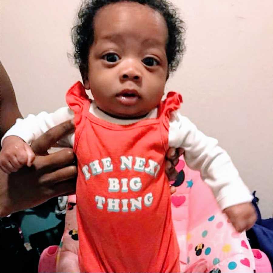 A baby girl with curly hair wearing a red outfit