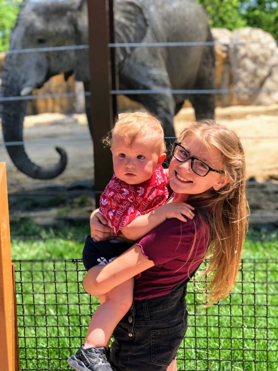 Sister holding baby sibling standing in front of elephant enclosure at zoo