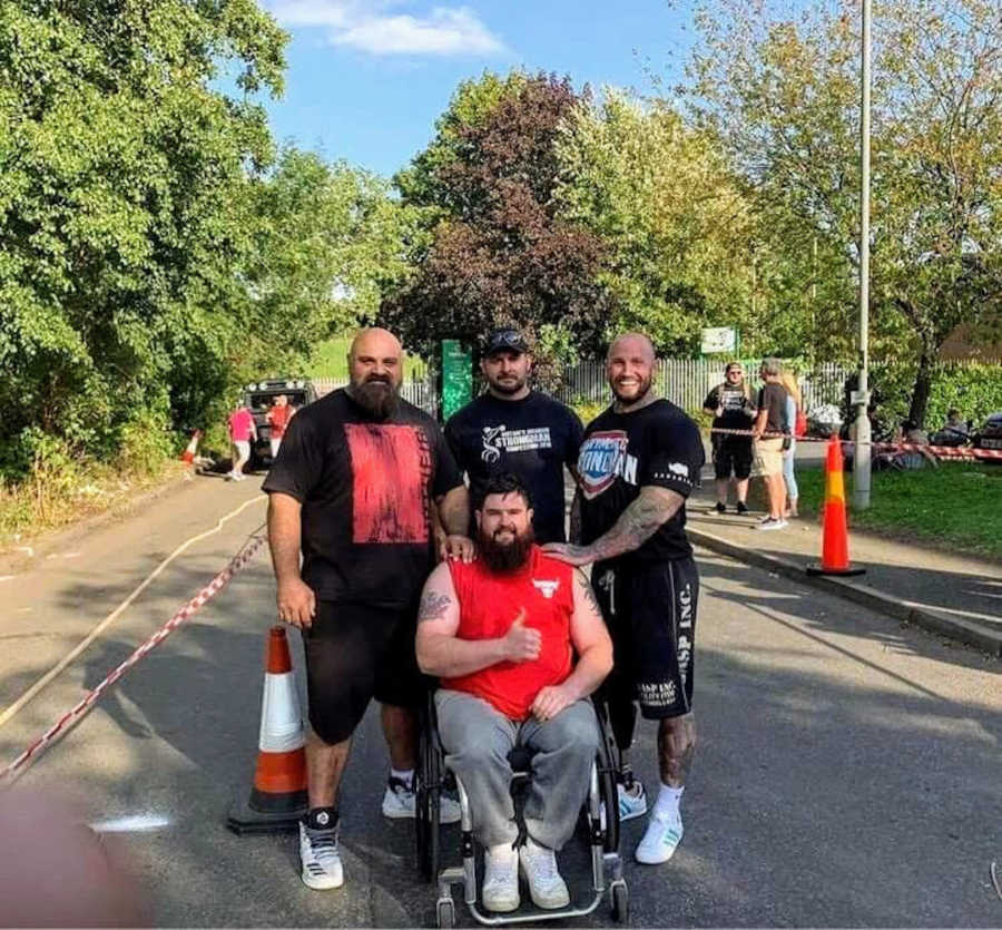 Three men standing behind man in wheelchair doing thumbs-up