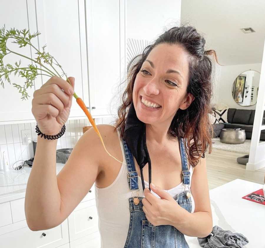 Woman in kitchen wearing overalls holding tiny carrot