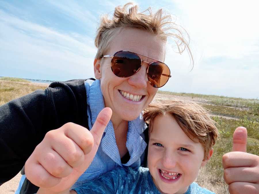 An autistic boy gives thumbs up to the camera while his mom smiles