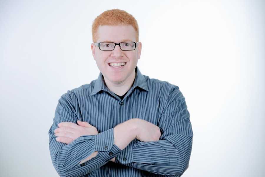 A stand-up comedian with red hair wearing a blue button-up and glasses