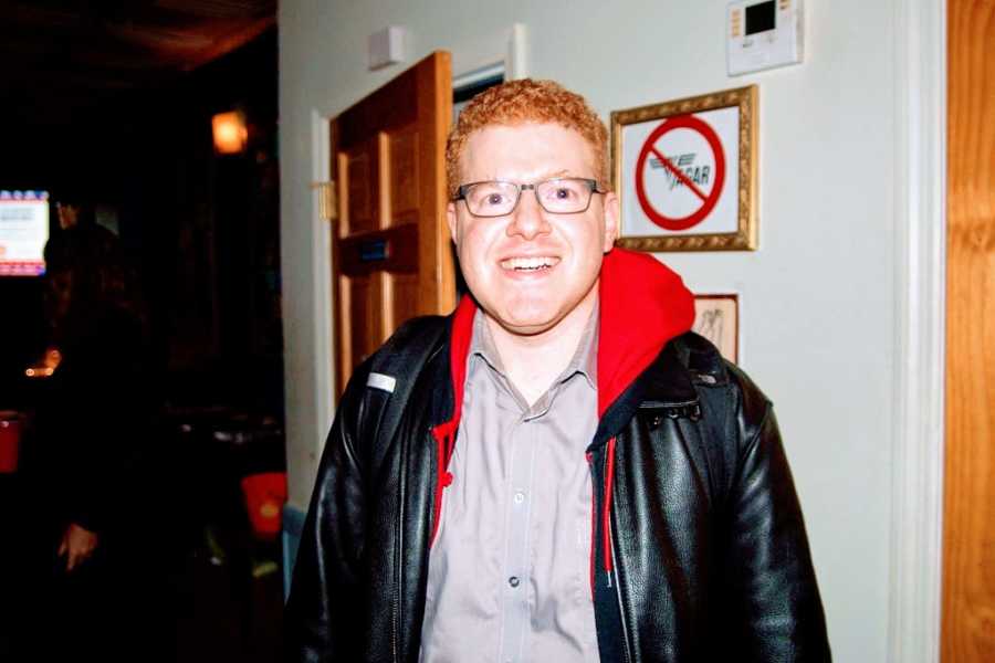 A comedian with red hair and glasses wearing a black leather jacket