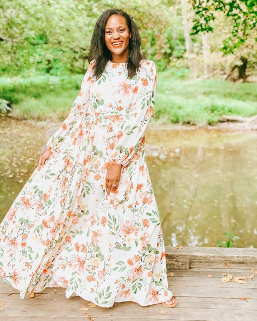 A woman wearing a floral dress stands on a bridge near a field