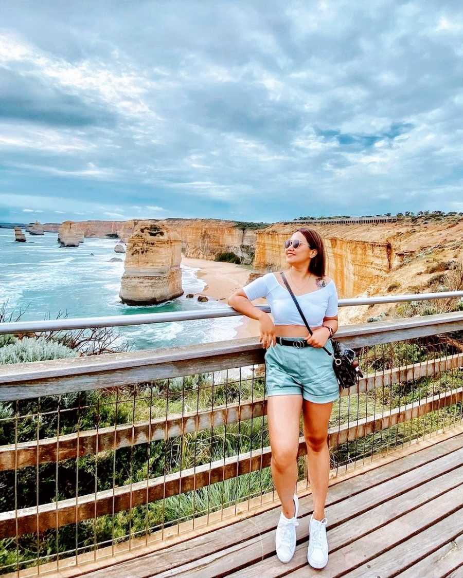 A woman stands on a bridge in front of cliffs and water