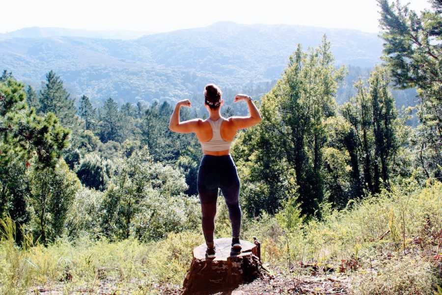 Woman flexing standing on a rock with back to camera in front of pine tree forest