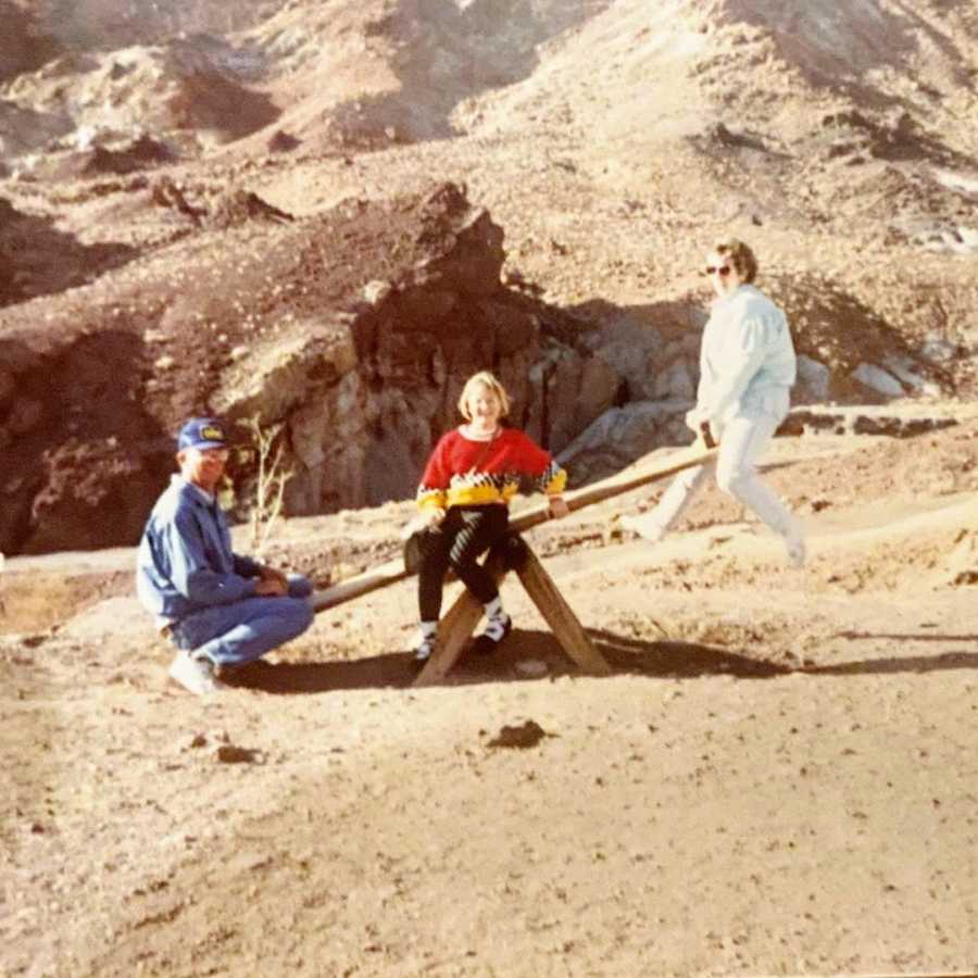 Mother and father sitting on teeter totter in desert with daughter in the middle