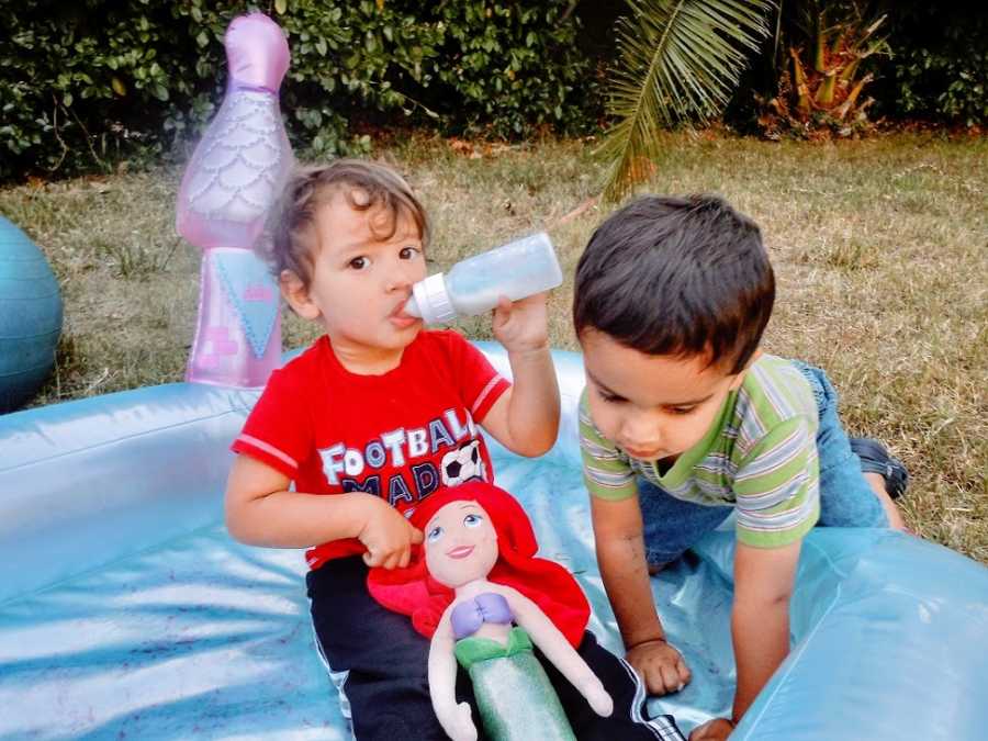 A boy sucks on a bottle while playing with a friend and a Little Mermaid doll