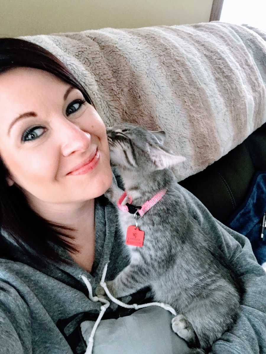 A woman sits on her couch with a kitten licking her face