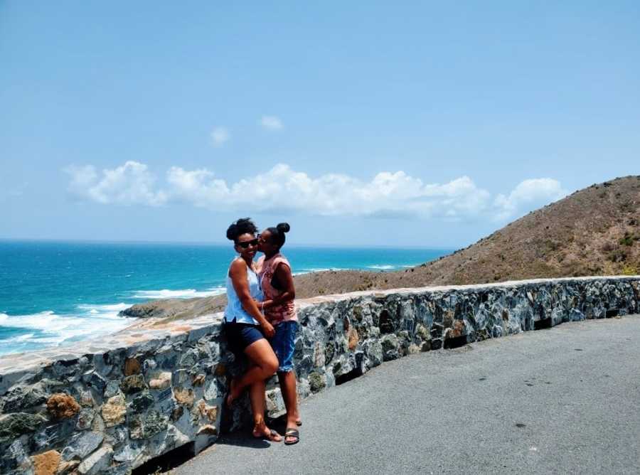 A woman kisses her wife on a road by the water