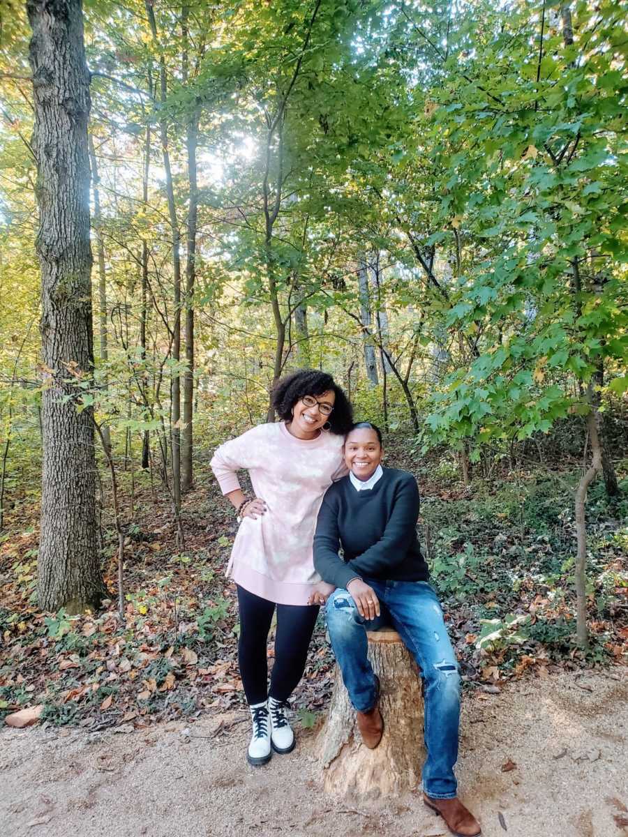 A woman sits on a stump while her wife leans on her shoulder in the woods