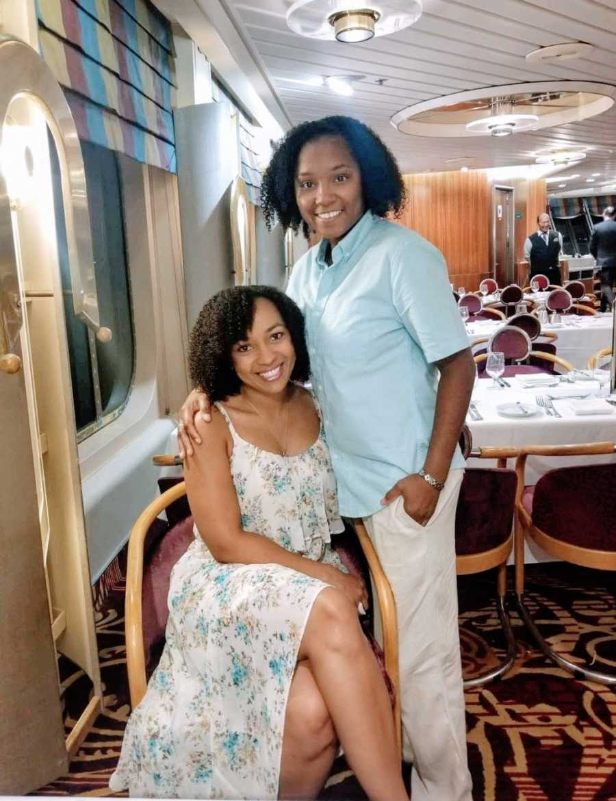 A Black woman sits in a chair while her wife stands with her arm around her