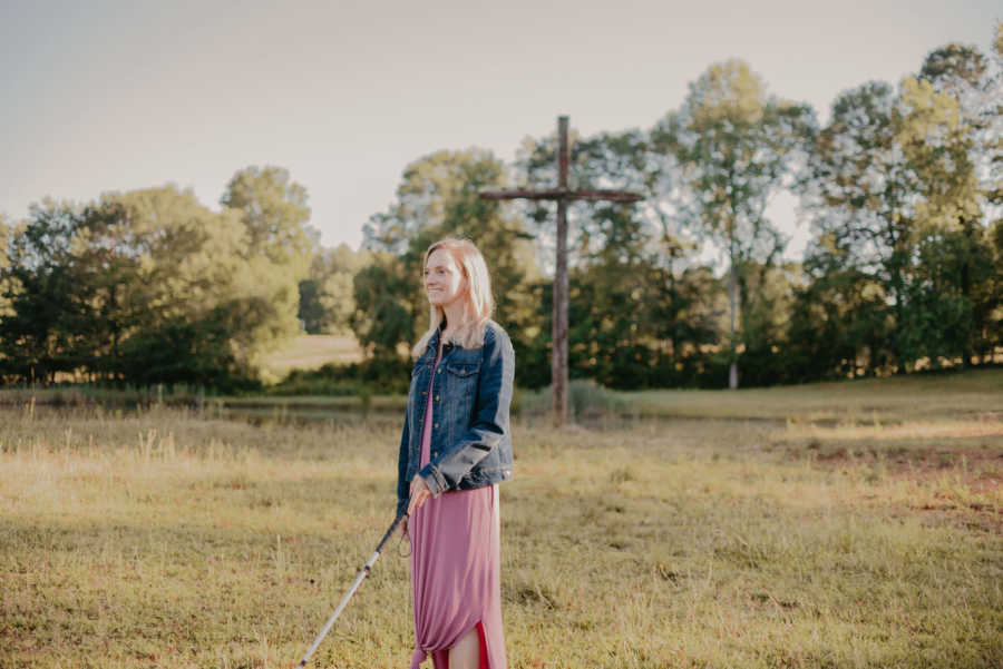 Blind woman using walking cane standing in field with wooden cross