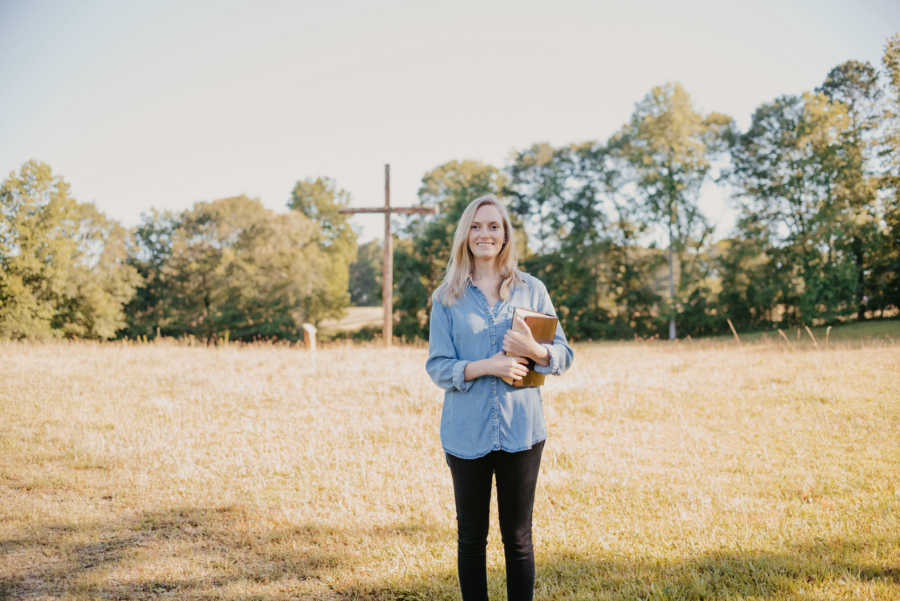 Woman standing in field with cross in background holding a Bible