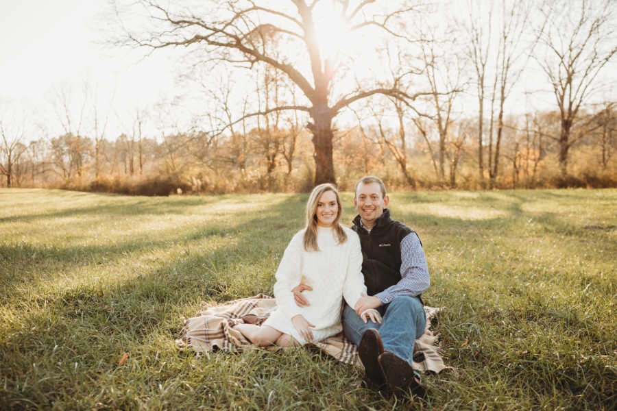 Husband and wife sitting on blanket in grassy area with trees