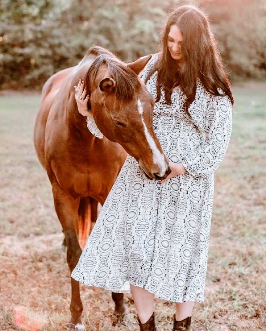 A pregnant woman stands with a brown horse