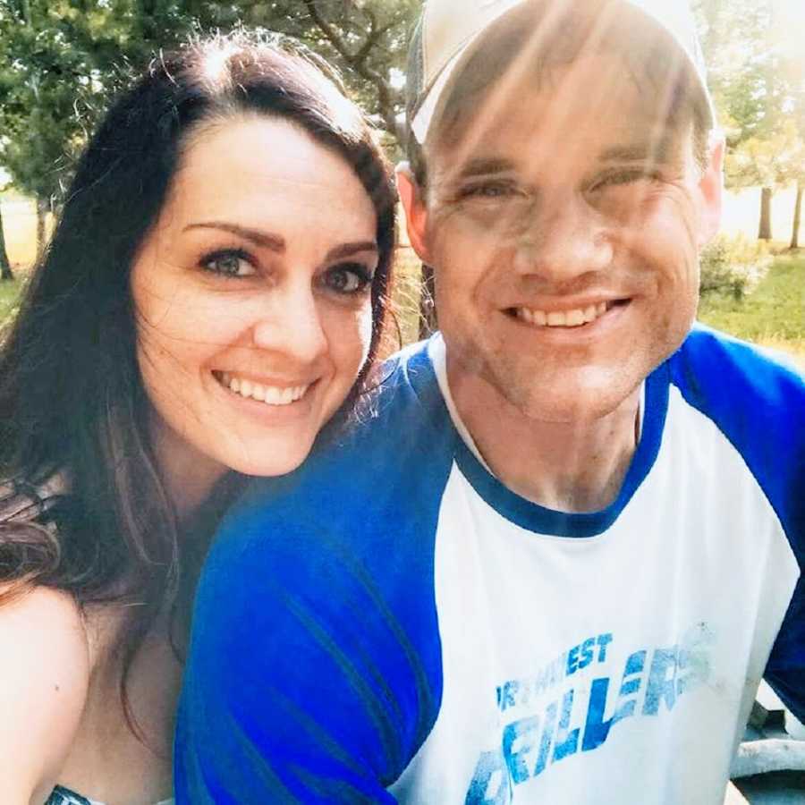 A woman sits with her husband outside, leaning her chin on his shoulder