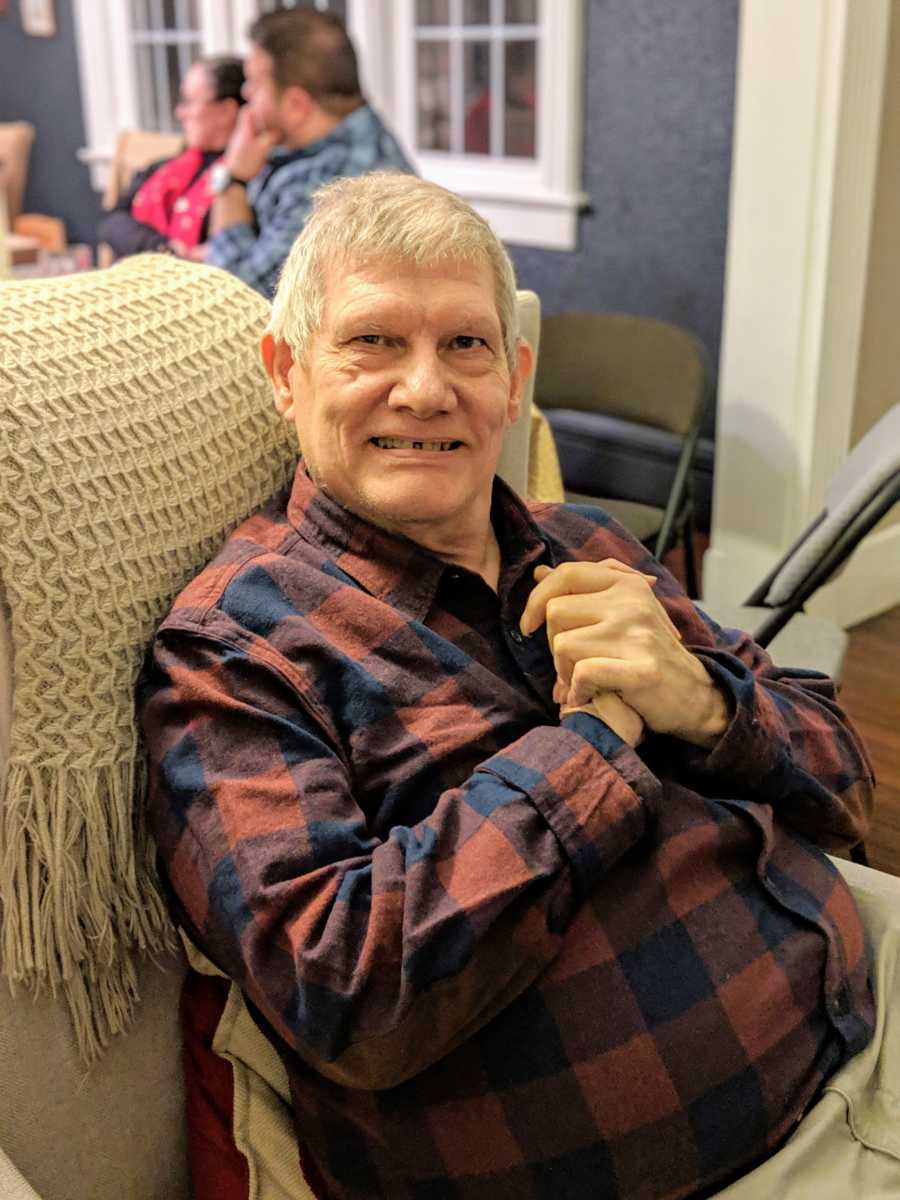 Older man sitting on beige couch with hands clasped on chest