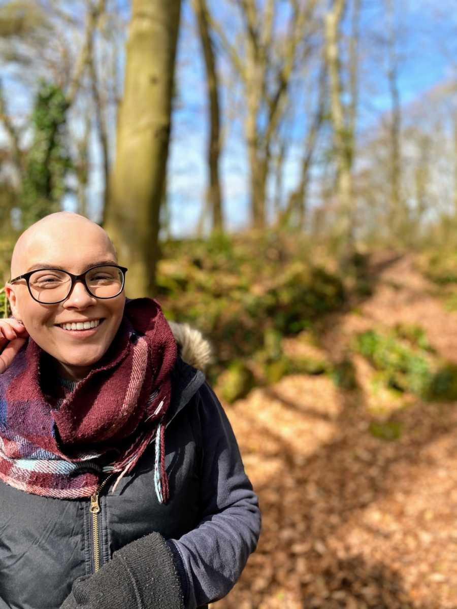 Bald woman wearing scarf and glasses standing in woods