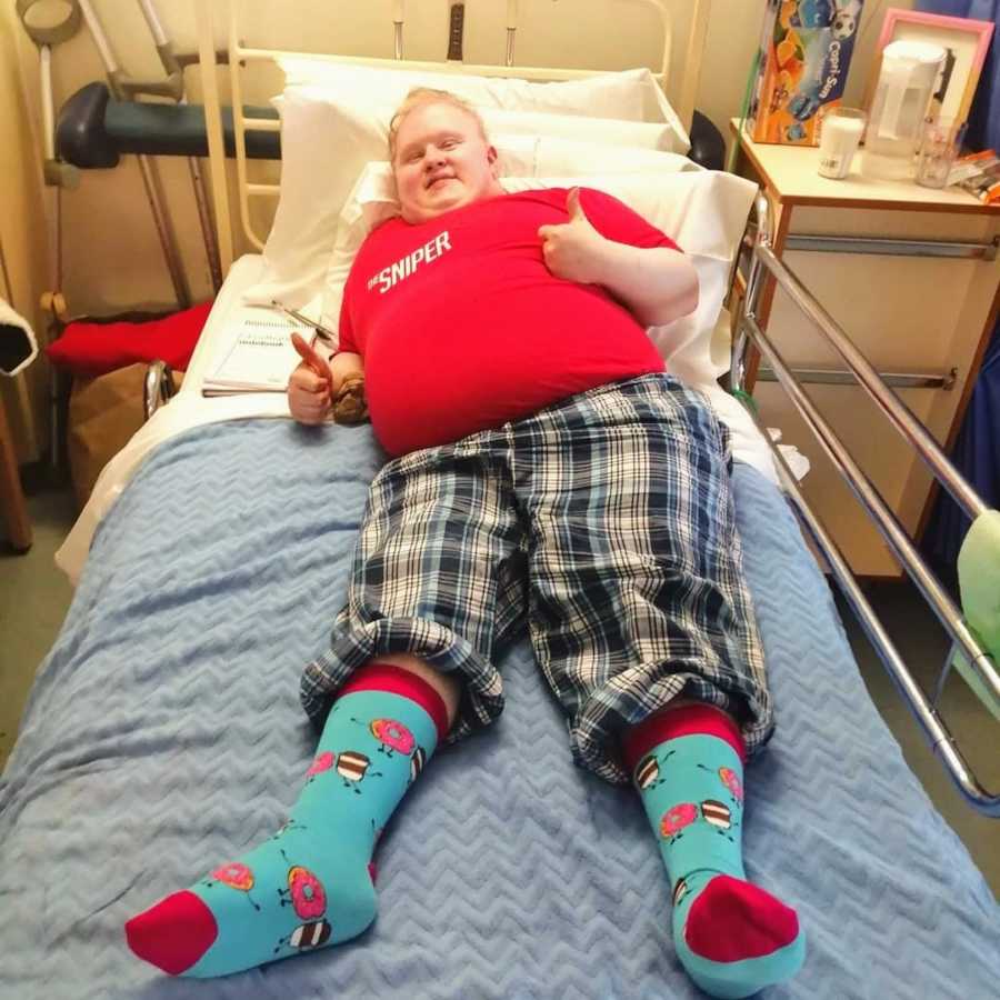 Young man lying in hospital bed wearing patterned socks giving thumbs up