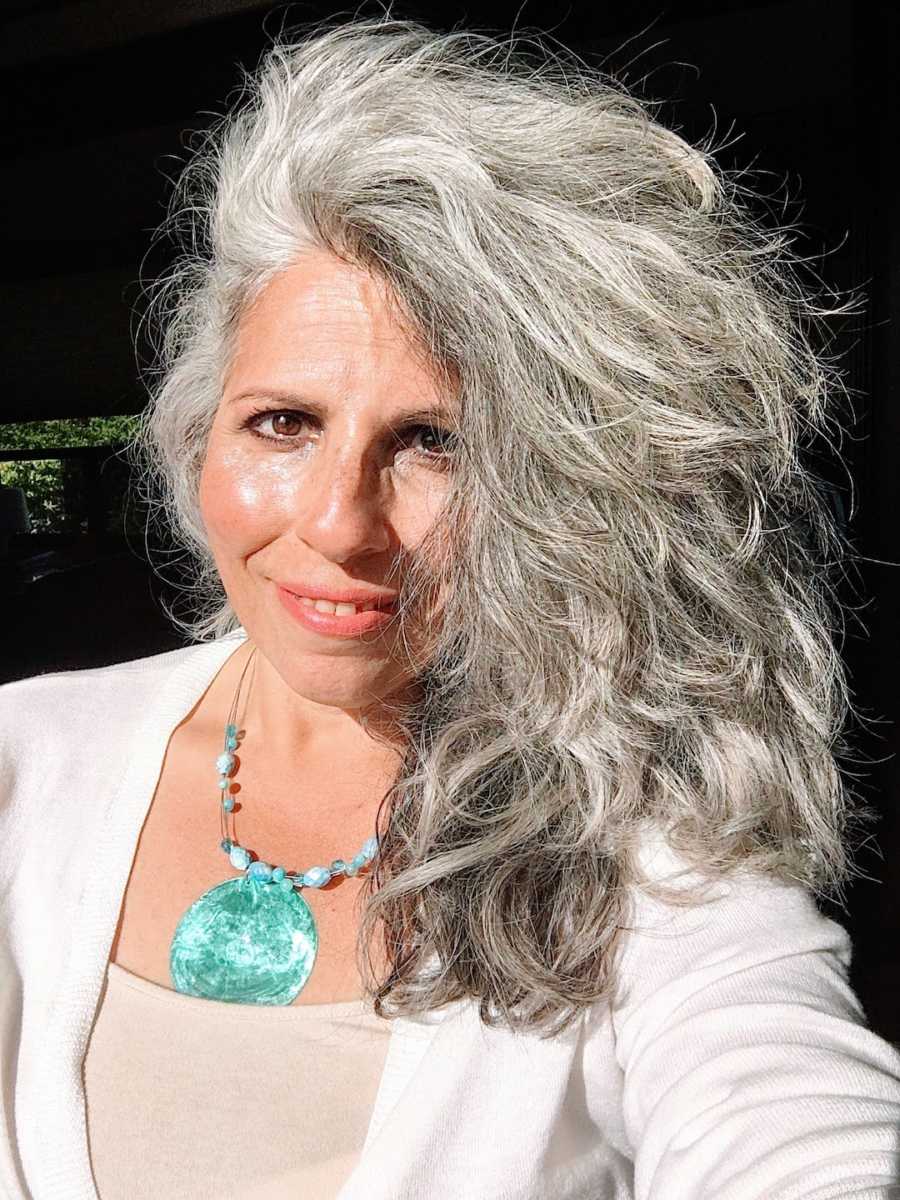 Woman with gray hair wearing blue shell necklace taking smiling selfie in the sunlight