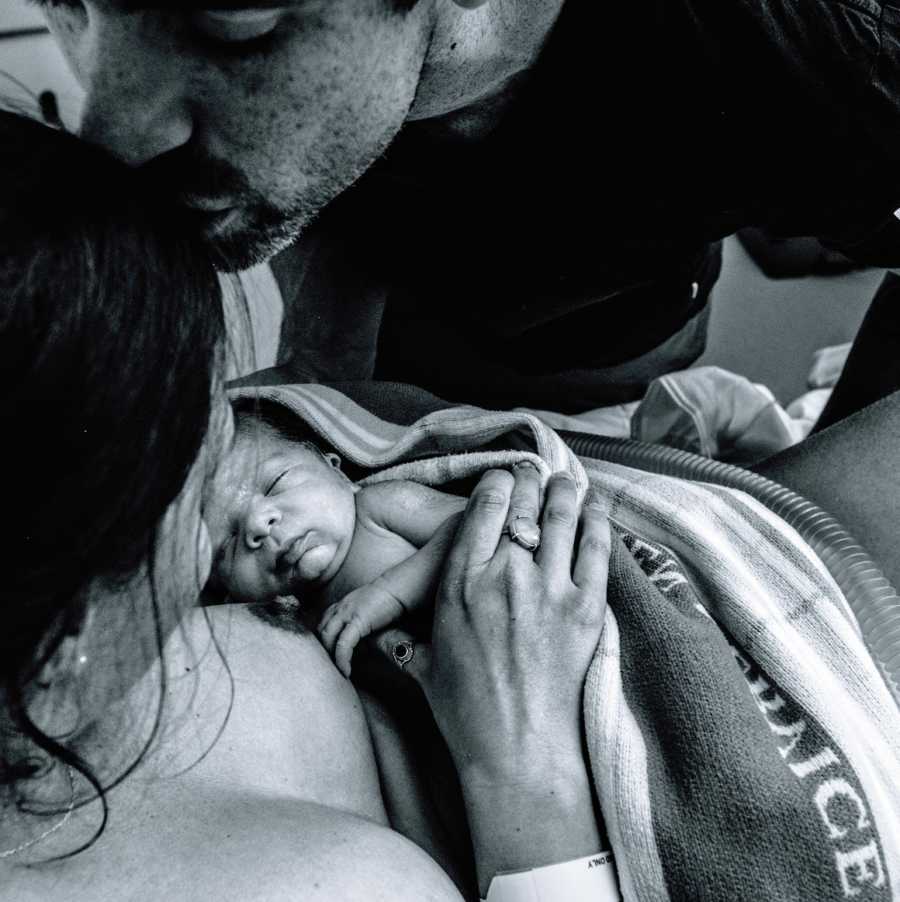 A mother holds her newborn rainbow baby while her husband kisses her forehead