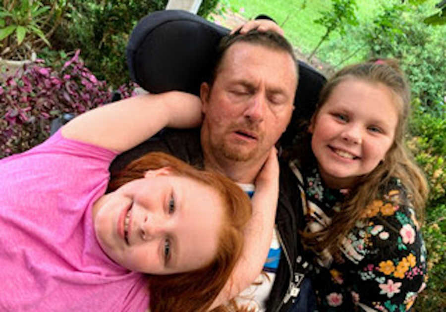 Father in wheelchair being hugged by two daughters