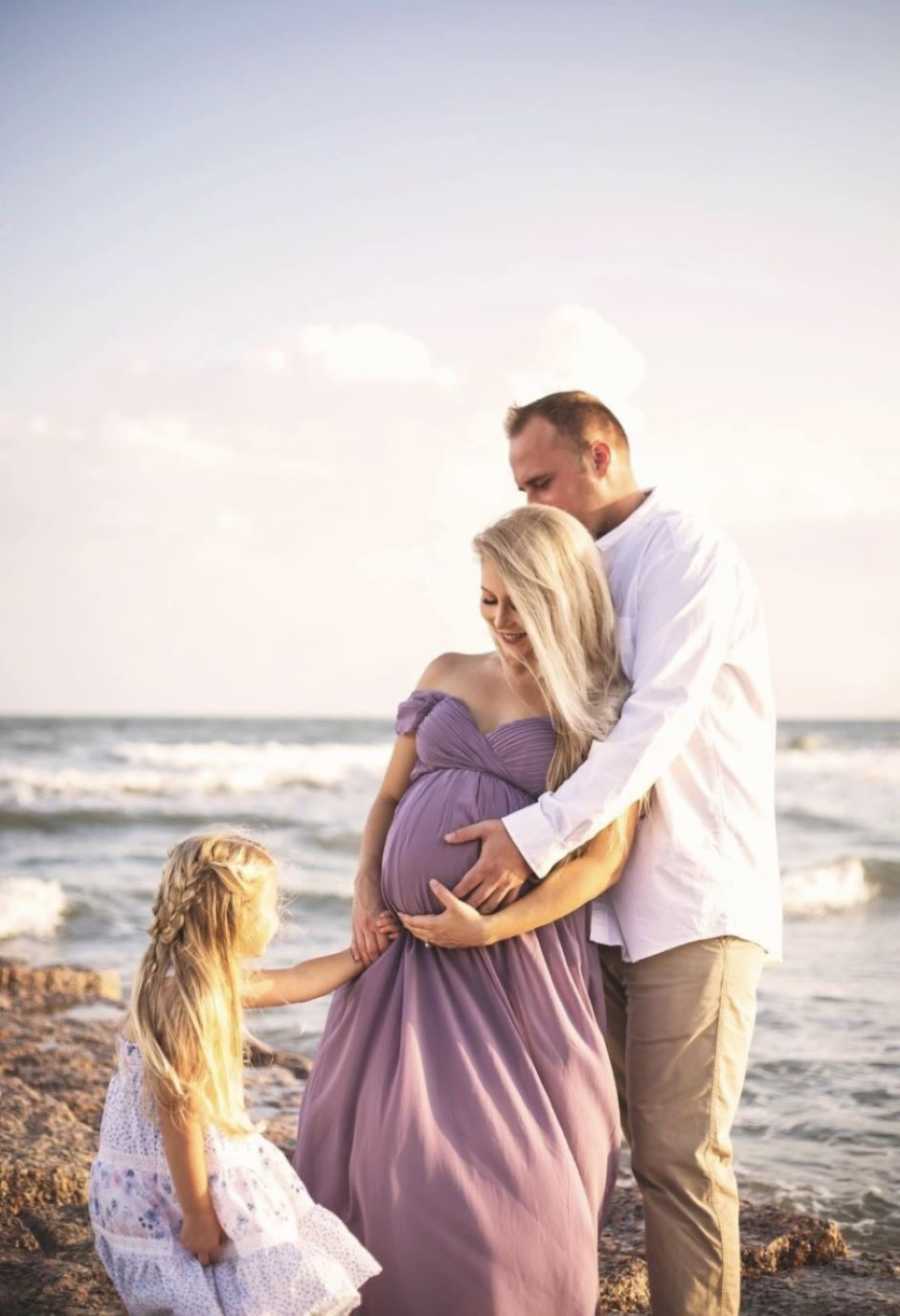 During maternity photoshoot on the beach, husband and daughter both touch mom's pregnant belly