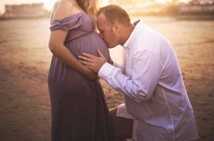 Husband kisses wife's baby bump during pregnancy photoshoot on the beach