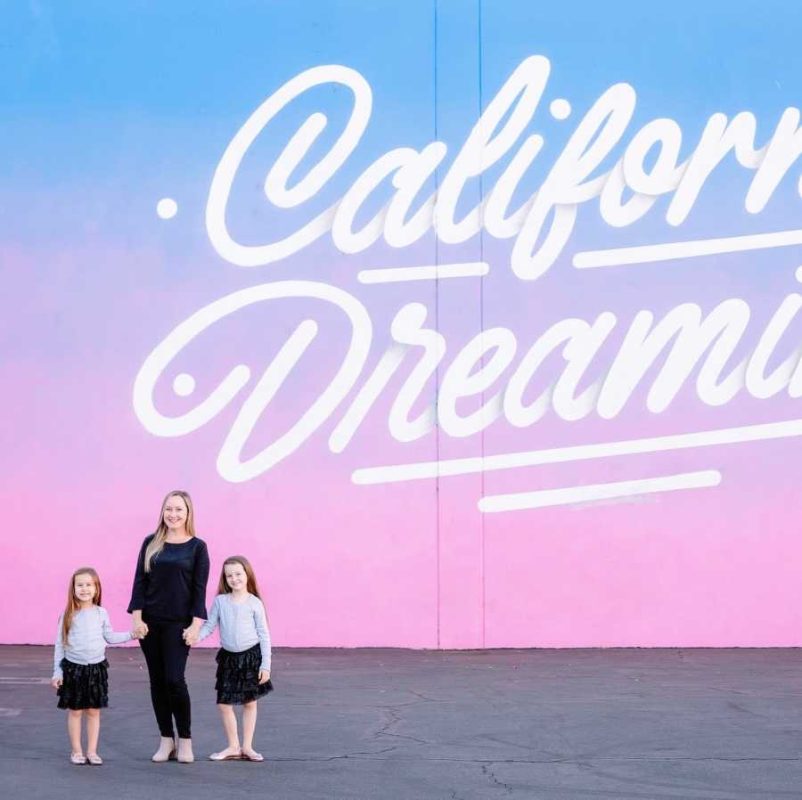 Mom stands in front of wall mural that reads "California Dreaming" with her two daughters in matching outfits