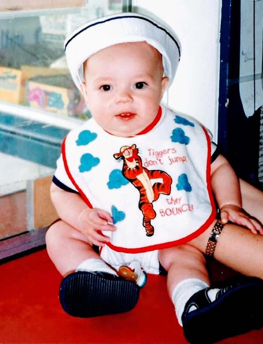 Mom takes photo of young son sitting near a window wearing a hat and a bib with Tigger on it