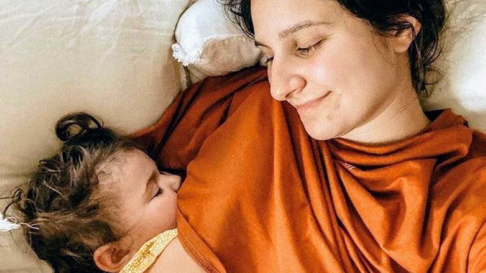 Young mom smiles down at her toddler while she sleeps next to her during an afternoon nap