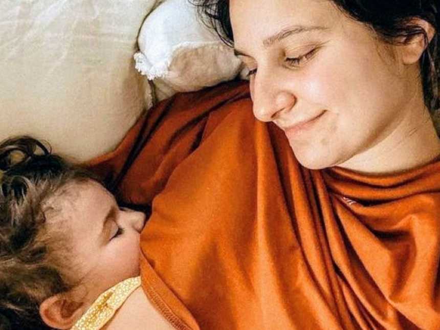 Young mom smiles down at her toddler while she sleeps next to her during an afternoon nap