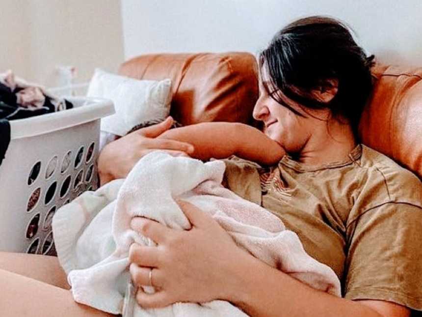 两个孩子的年轻妈妈在沙发上给女儿喂奶，旁边放着一堆要洗的衣服