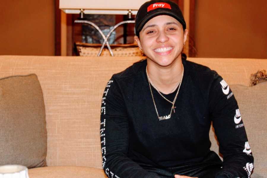 Young lesbian smiles for the camera dressed in masculine clothing and a baseball cap that says 'pray'