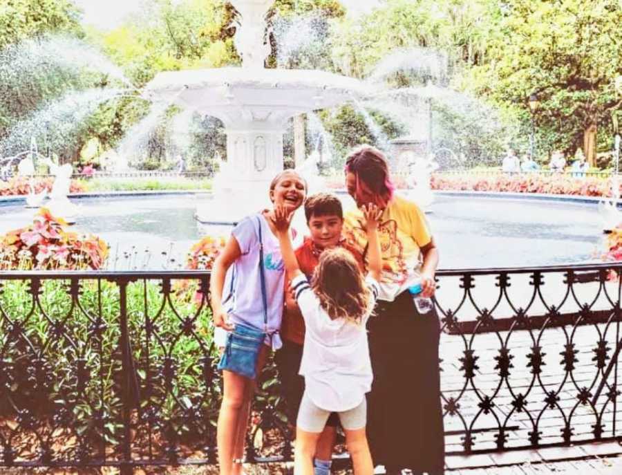 Mom smiles down at her three kids while posing for a photo on vacation in front of a fountain