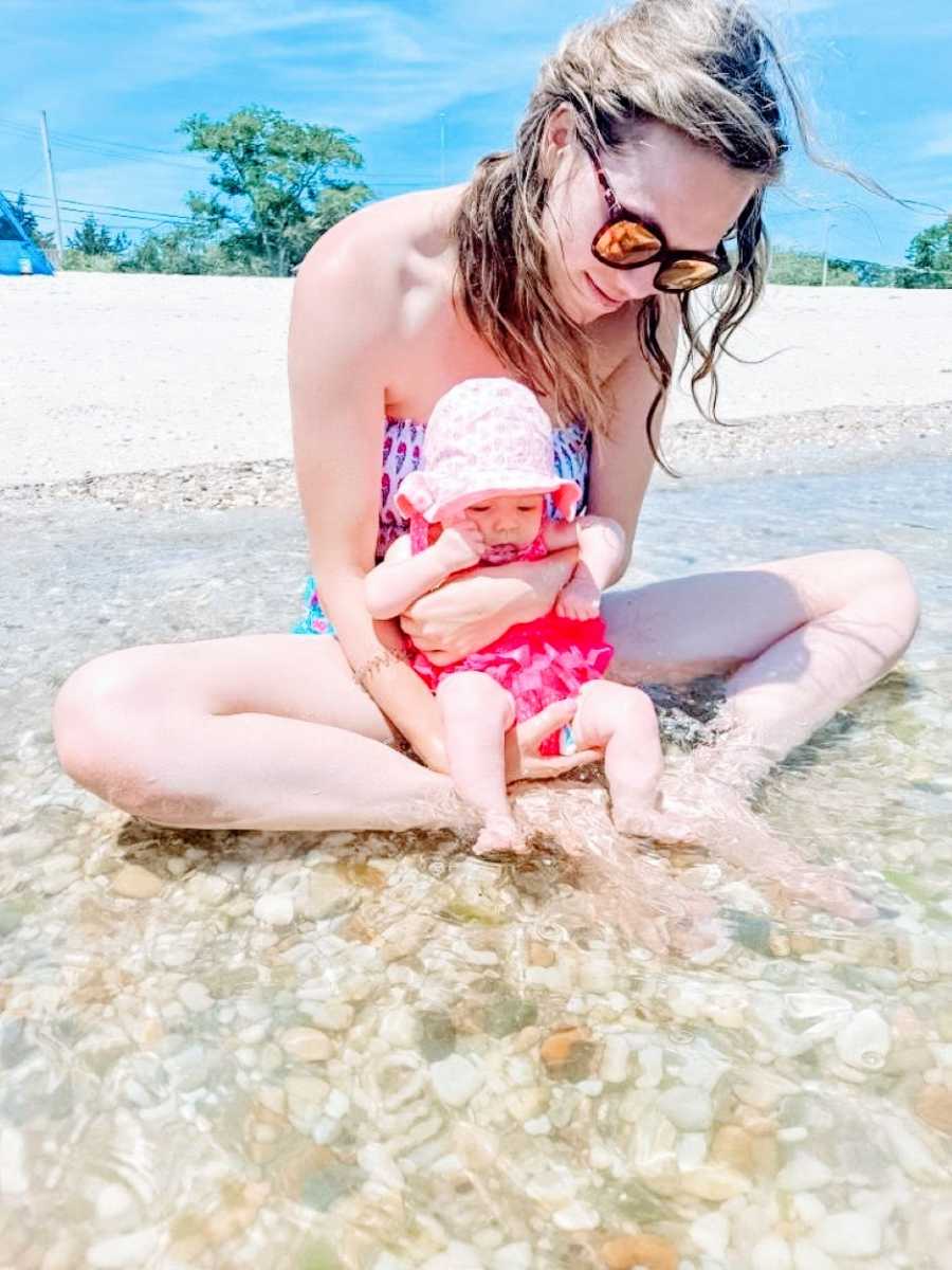 Girl mom takes photo with newborn water while they sit in calm waters at the beach