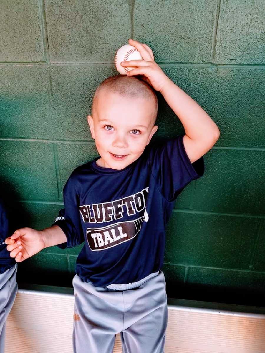 A little boy holds a baseball on top of his head by a green wall