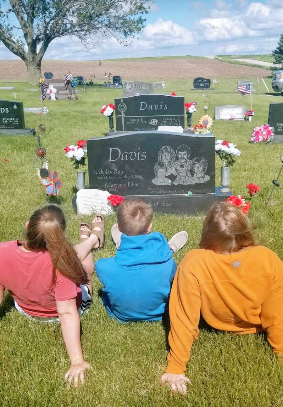 Three children sit in a graveyard looking at a headstone