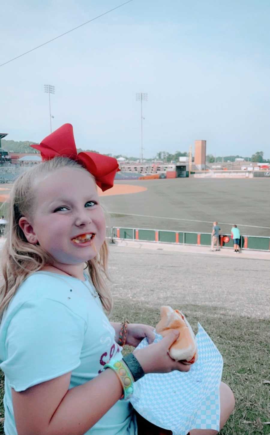 A young girl wearing a bow smiles with foot in her mouth