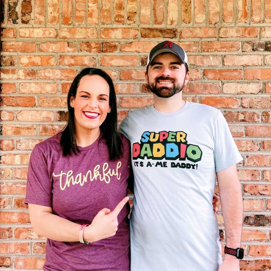 A woman points to her husband who has his arm around her