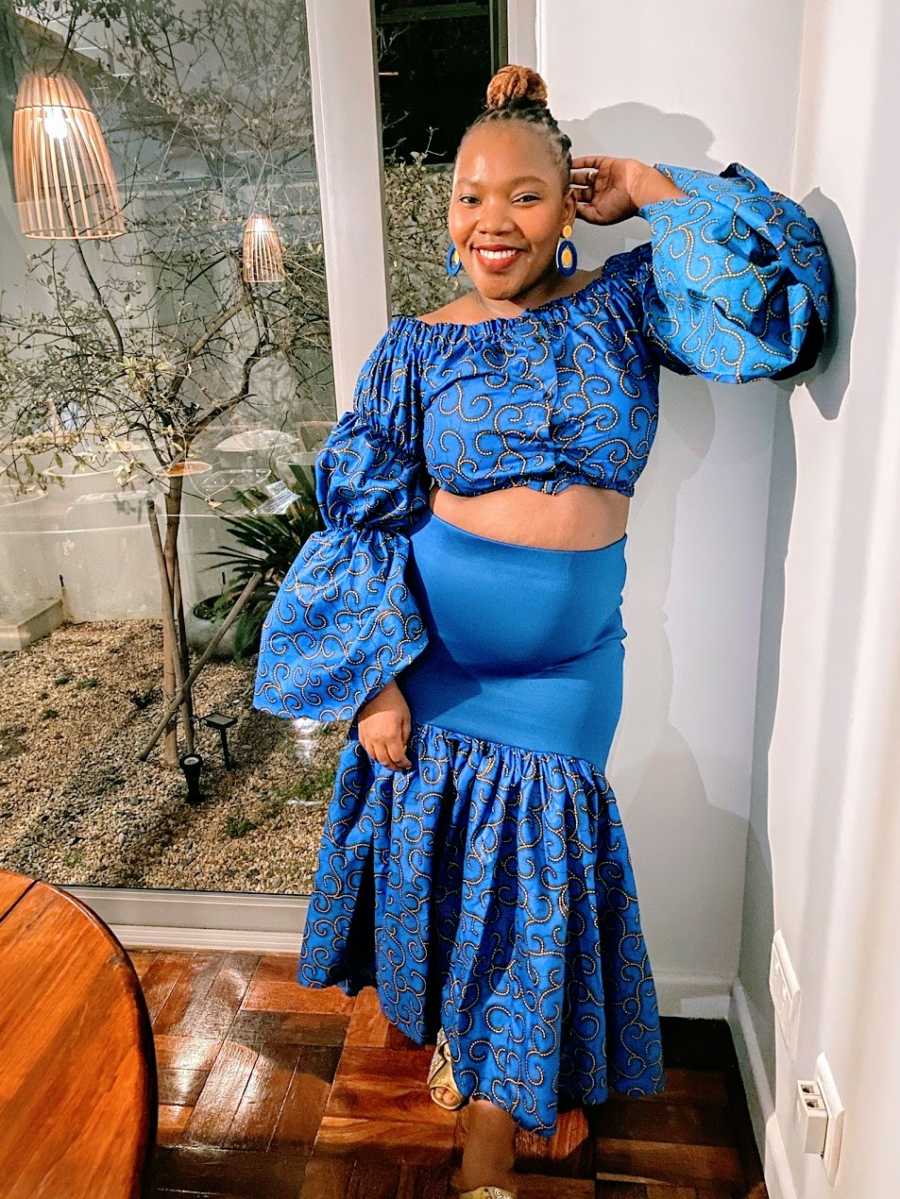 A woman wearing a blue dress and blouse leans against a wall