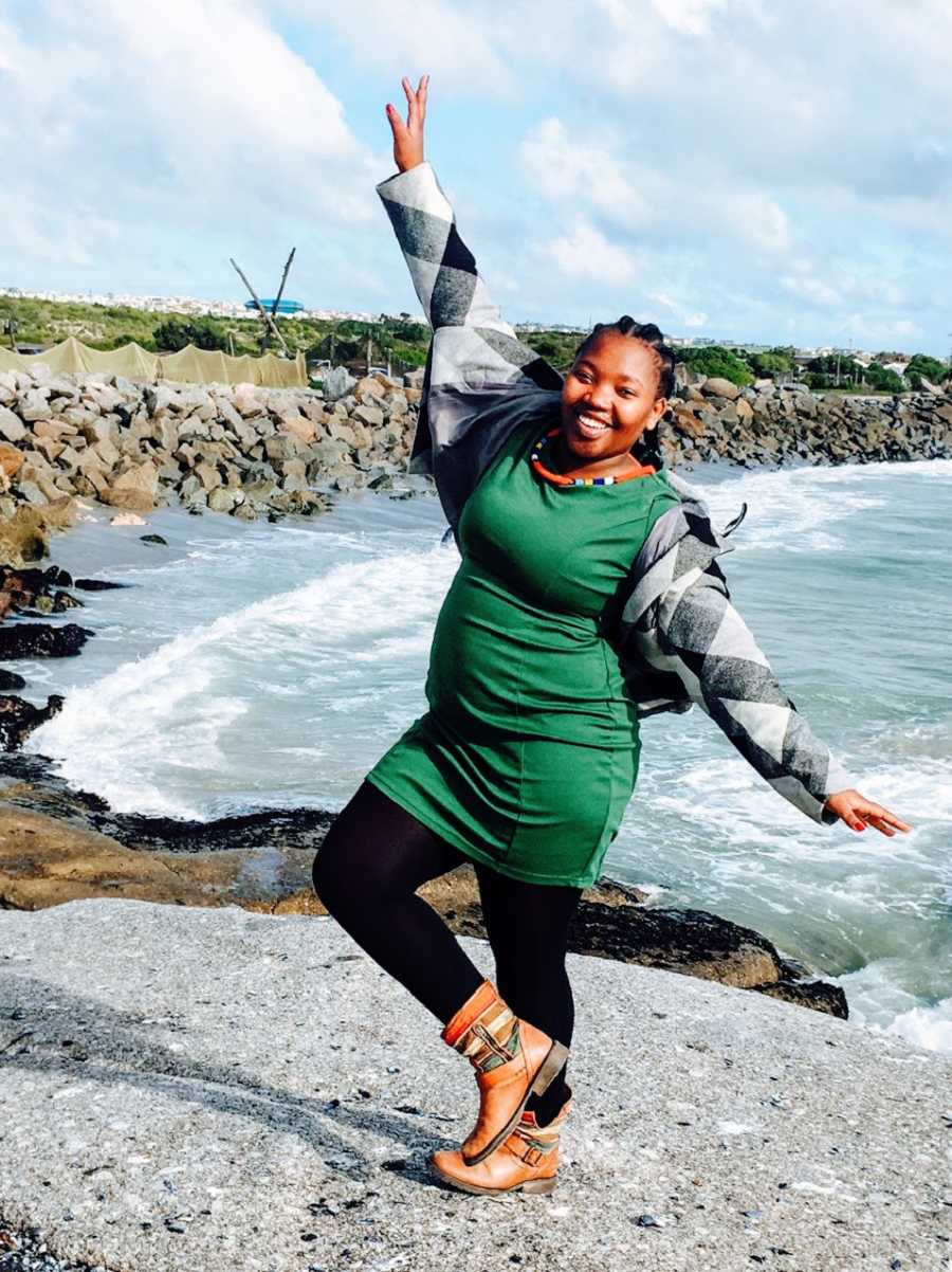 A woman wearing a green dress and a checkered jacket stands on one leg by the water