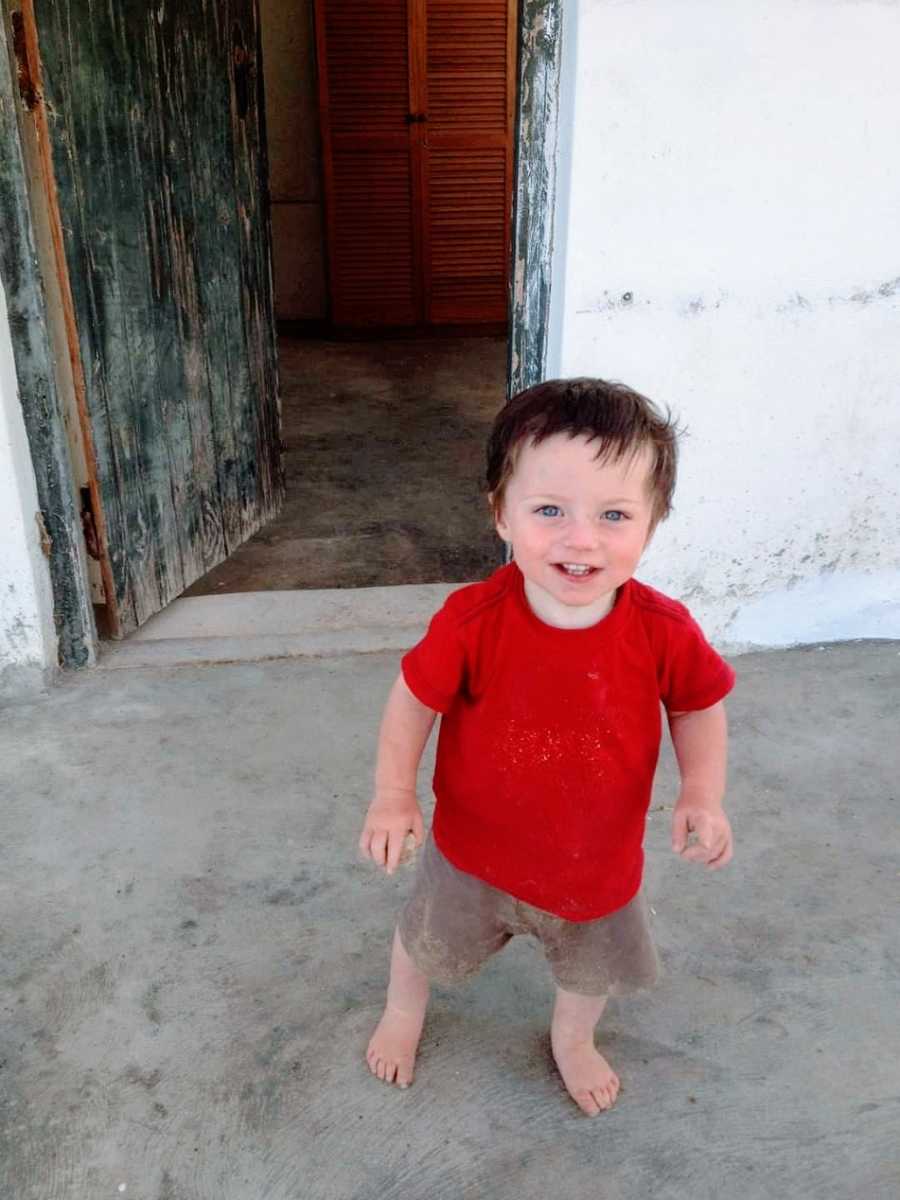 A boy in a red shirt and shorts stands outside a door
