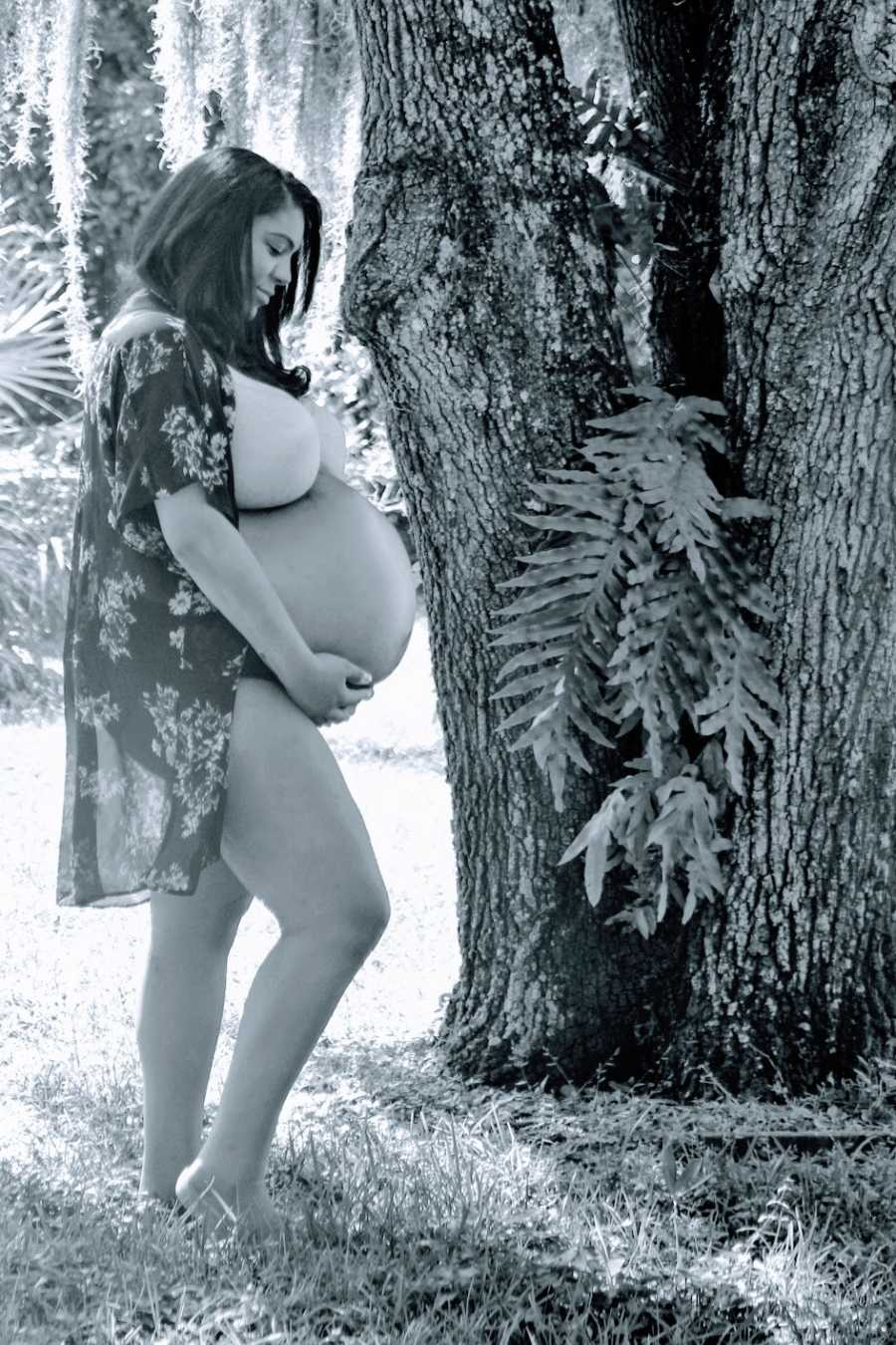 A pregnant woman holds her stomach outside by a tree