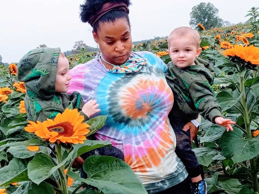 A mother holds her twin sons walking through a field of sunflowers