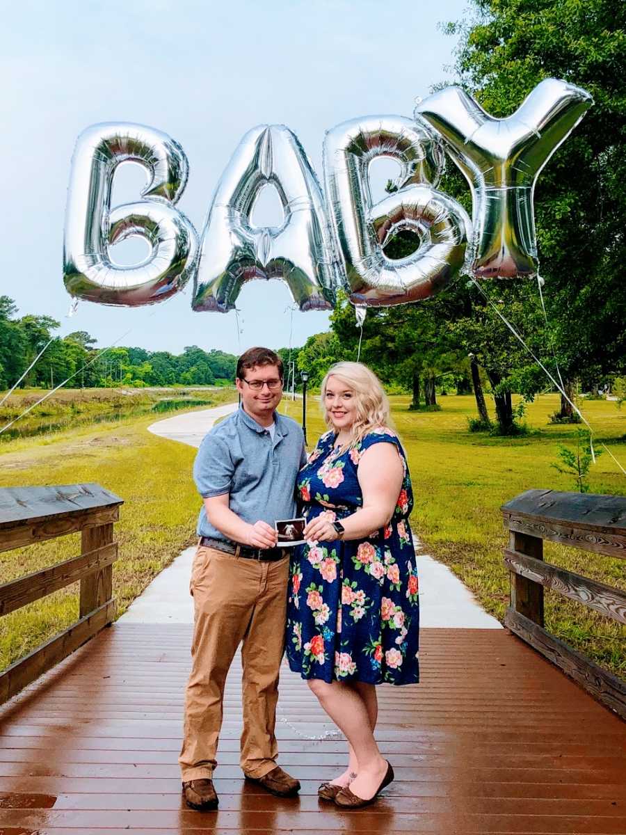 A woman and her husband stand under balloons spelling BABY