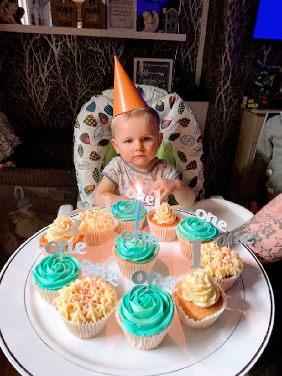 A little boy wearing a birthday hat sits in front of cupcakes