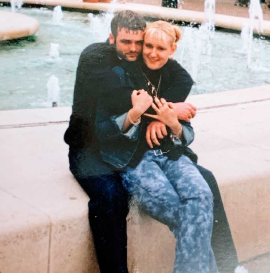A woman sits on her husband's lap by a water fountain